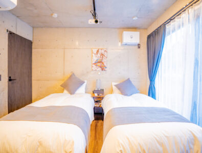 Modern twin bedroom at Bayside Villa Miyako Island with white bedding, blue curtains, wooden floors, projector, and contemporary amenities.